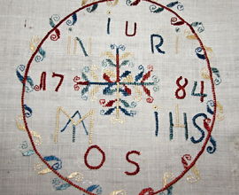 Křestní šátek, bílé plátno, barevně vyšívaný hedvábím, motiv kytice, věneček s monogramy a letopočet „1784“. Darovala paní Josefa Náprstková v roce 1887. H4-NČ -1527